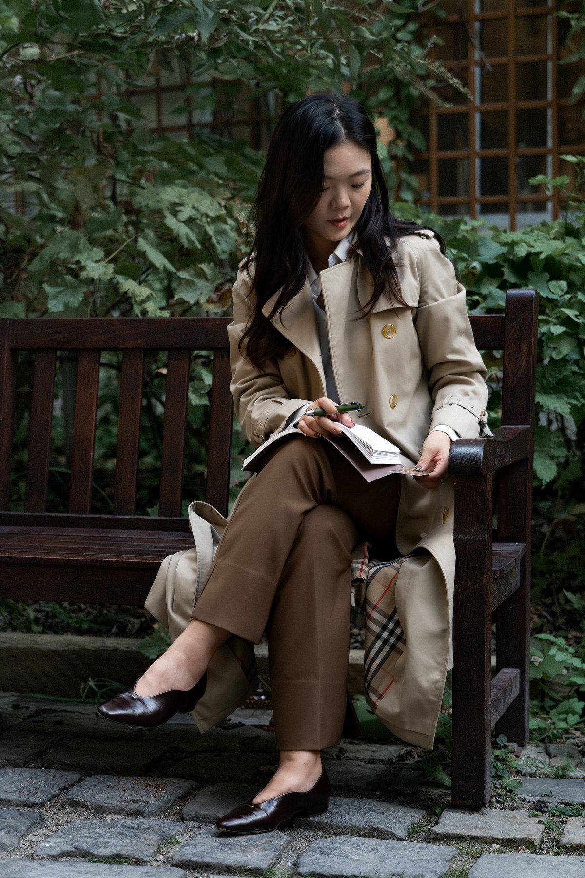 Nina Xu on a garden bench in a trench coat, writing in a notebook